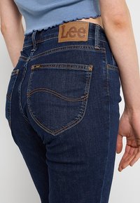 Jean en denim bleu foncé avec surpiqûres orange contrastantes, détails sur la poche arrière et un patch logo Lee marron sur la ceinture porté avec un haut court bleu.