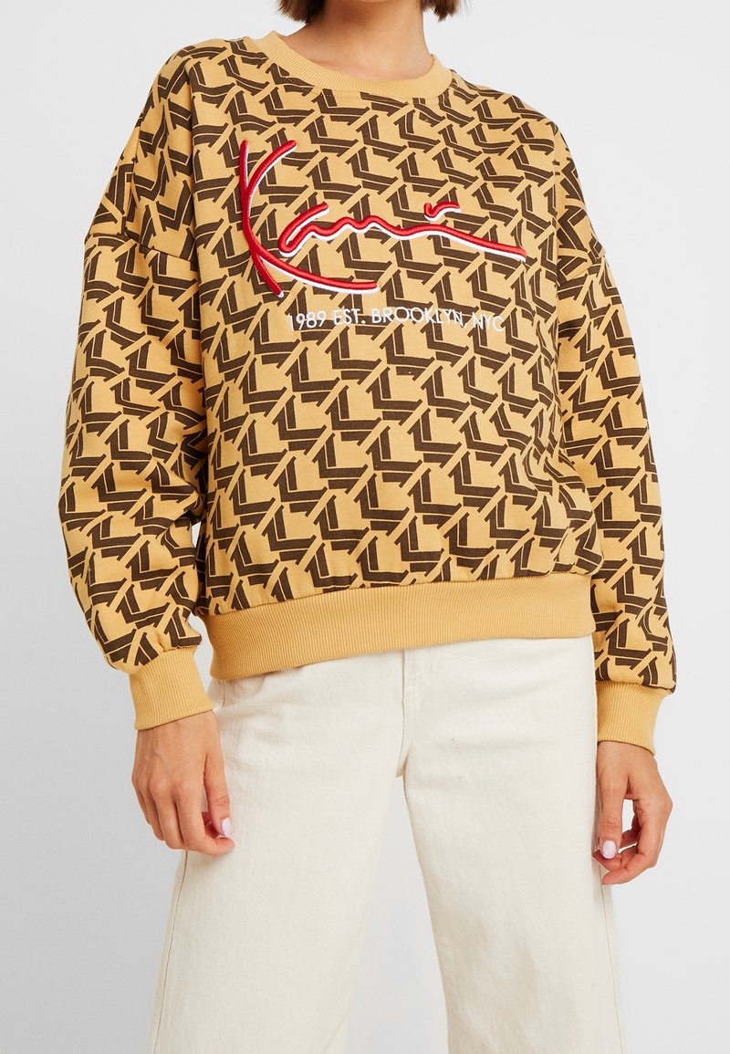 Sudadera color mostaza con un patrón geométrico oscuro, texto bordado en rojo "Kani" y texto en blanco "1989 Est. Brooklyn NYC", combinada con pantalones blancos.