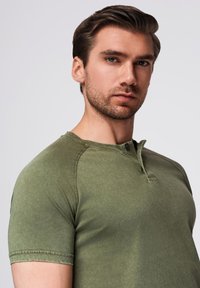 Olivově zelené tričko s krátkým rukávem typu henley ze měkké látky s texturovaným povrchem, se zapínáním na knoflíky a raglánovými rukávy.