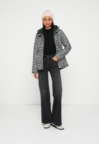 Μπροστά μαύρο-άσπρο μοτίβο μπουφάν χειμερινό με κουκούλα, συνδυασμένο με μαύρο πουλόβερ και σκουρόχρωμο τζιν καμπάνα. Το μοντέλο φοράει ροζ σκούφο.