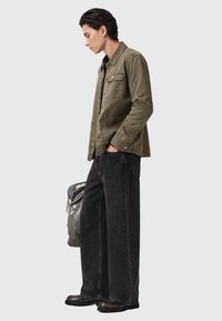 Giacca verde oliva con dettagli in bottone, abbinata a jeans neri larghi in denim. Stivali neri resistenti e una borsa grigia scura completano il look.