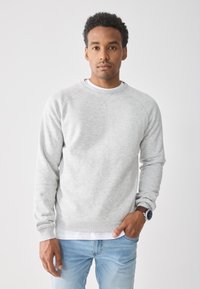 Joven con cabello corto y rizado, vistiendo un sudadera gris claro, camiseta blanca debajo, jeans azules y un reloj, de pie contra un fondo sencillo.