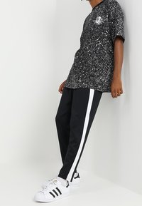 Osoba v černém tričku s tečkami, černých kalhotách s bílými bočními pruhy a bílých teniskách s černými pruhy se opírá o bílou zeď.