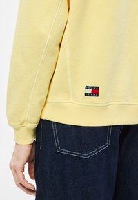 Κίτρινο φούτερ με ριμπ μανσέτες και τελείωμα, που διαθ�έτει μια μικρή ετικέτα με λογότυπο σε κόκκινο, λευκό και μπλε. Συνδυάζεται με σκουρόχρωμο τζιν παντελόνι.