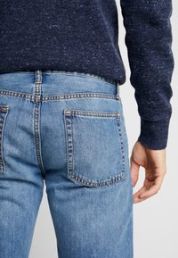 Person som bär ljusblå denimjeans och en mörkblå prickig tröja, med en hand avkopplad vid deras sida.