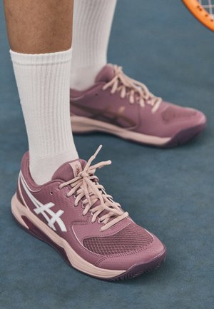 Zapatillas deportivas moradas con parte superior de malla, suela de goma sólida y acentos blancos. Combinadas con calcetines blancos de canalé, sobre una superficie azul.