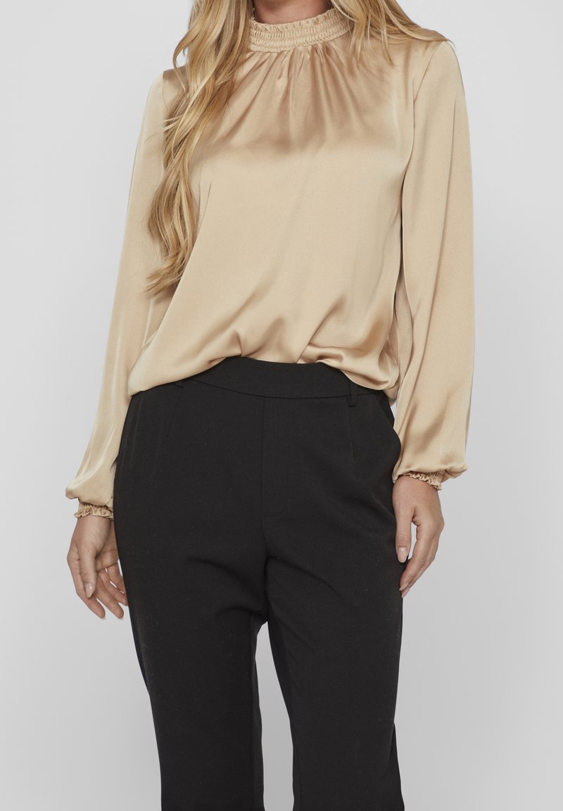 Femme portant un chemisier beige en satin à manches longues avec col à volants et un pantalon noir taille haute sur mesure, posant devant un fond gris.