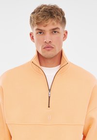 Lichtoranje rits sweatshirt met een hoge kraag, gestikte details en een logoaccent. Zachte stoftextuur met geribbelde manchetten.