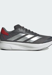 Zapatilla deportiva gris con una parte superior de malla texturizada, tres franjas plateadas, acentos rojos en el talón y una entresuela blanca ligera.