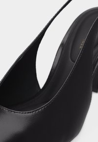 Μαύρα δερμάτινα γυναικεία παπούτσια με λουράκι πίσω, ψηλοτάκουνα με μυτερά δάχτυλα, ορατή ραφή και λεία υφή σε φωτεινό φόντο.