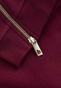 Burgundská mikina z textílie s rebrovanými manžetami. Má zlatý zips s obdĺžnikovým úchytom so zalisovaným logom. Hladká textúra.