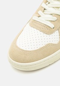 Beige mockaskor med vit perforerad läderpanel, med snören av flata skosnören och strukturerad gummisula. Rundad tå med stickningsdetaljer.