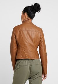 Hnědá kožená bunda s přiléhavým střihem, polstrovanými detaily na ramenou a zkrácenou délkou. Nosená přes zelené kalhoty.