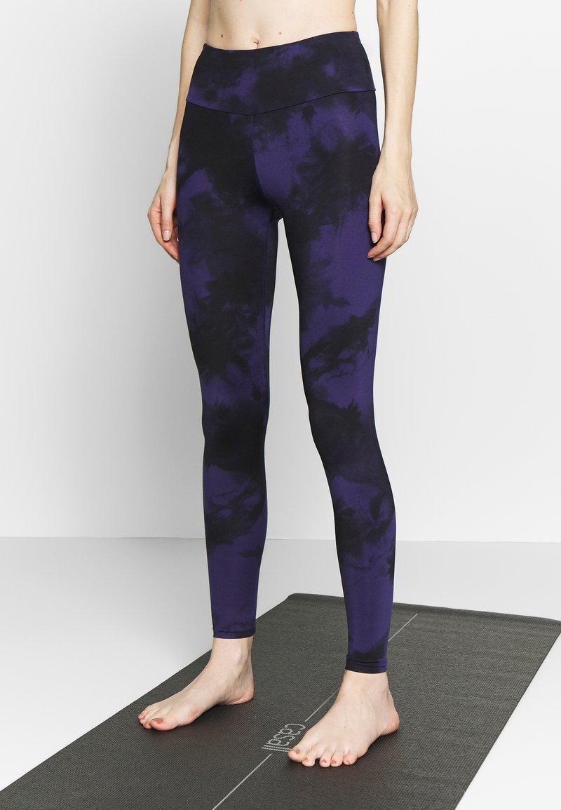 Fioletowe legginsy tie-dye z wysokim stanem i gładką fakturą, noszone na boso, na ciemnej macie do jogi.