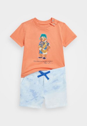 Oranges Baumwoll-T-Shirt mit einem Bärenmotiv und einem blauen Bucket Hat-Design, kombiniert mit hellblauen Batik-Shorts mit blauer Bindeschnur.