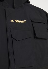 Veste imperméable noire avec le logo Adidas Terrex en or. Elle dispose de plusieurs poches frontales avec des fermetures à rabat et d'un tissu texturé.