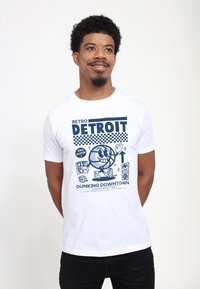 Bílý tričko s retro modrým grafickým designem obsahující nápis "Detroit", basketbalovou tématiku a ilustrace, včetně hráče a ikon.