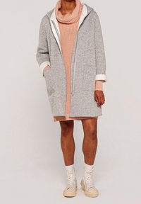 Szary, dzianinowy, oversize'owy płaszcz z białymi wykończeniami, kieszeniami z przodu i z zapięciem na zamek, noszony na różowej sukience o prążkowanej strukturze. W komplecie z kremowymi, wysokimi sneakersami.
