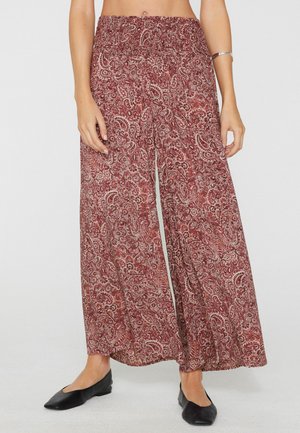 Donna che indossa pantaloni larghi e morbidi con fantasia paisley rossa e bianca e ballerine nere, in piedi davanti a uno sfondo uniforme.