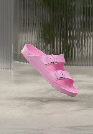 Sandale rose à bout ouvert avec deux sangles réglables à boucle, semelle texturée, flottant au-dessus d'un sol en béton lisse et d'un fond en verre ondulé.