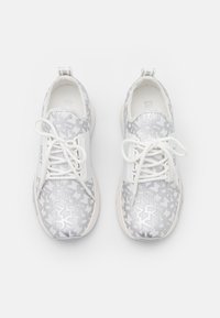 Ein Paar weiße Sneaker mit grauem "DKNY"-Logodruck, weißen Schnürsenkeln, die zu Schleifen gebunden sind, und weißen Sohlen, von oben auf einem einfarbigen Hintergrund betrachtet.