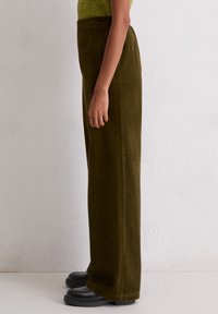 Maslinasto zelene kord pantalone ravnog kroja, s vertikalnom rebrastom teksturom i visokim strukom. Uparene s crnim mokasinama.