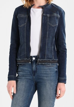 Kvinde iført en mørkeblå denimjakke over en hvid top kombineret med mediumblå denimjeans, stående med hænderne afslappet langs siderne.