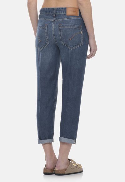 Personne portant un jean en denim bleu retroussé et des sandales beiges à enfiler avec de grandes boucles dorées, vue de dos sur fond blanc.