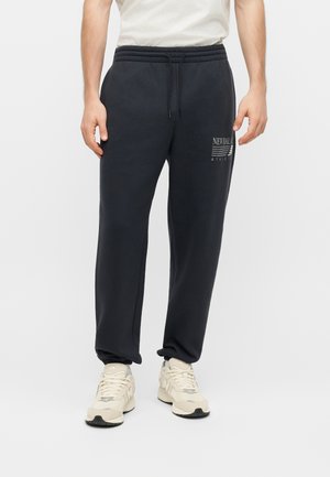 Hombre vestido con pantalones deportivos negros con el logo de New Balance en el muslo y zapatillas blancas, de pie frente a un fondo blanco liso.