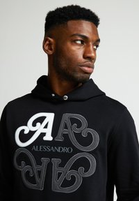 Sudadera negra con letras bordadas en relieve en blanco y gris "A" y "V" y la palabra "ALESSANDRO" en una fuente estilizada en la parte delantera.