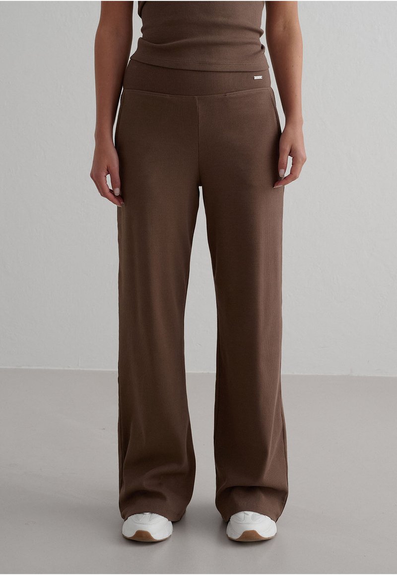 Personne portant un pantalon large marron et un haut assorti, debout sur un sol gris avec des baskets blanches.