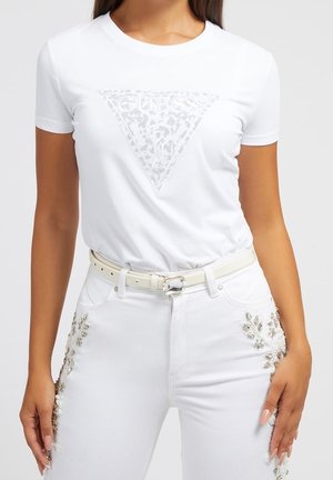 Kvinde iført en hvid Guess-logo t-shirt og hvide jeans med sølvfarvet blomsterbroderi samt et hvidt bælte med en klar spænde.