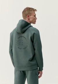 Zelený pulóver s kapucňou a textúrovaným logom na chrbte, vybavený šnúrkou na kapucni a rebrovanými manžetami. Mäkká tkanina, voľný strih.