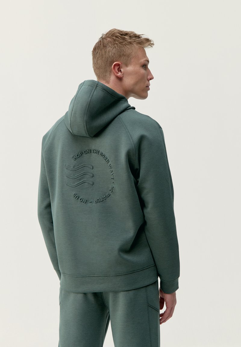 Zelený pulóver s kapucňou a textúrovaným logom na chrbte, vybavený šnúrkou na kapucni a rebrovanými manžetami. Mäkká tkanina, voľný strih.