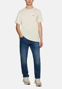 T-shirt crema a maniche corte con taschino, abbinata a jeans in denim blu. La maglietta presenta un col rotondo e un tessuto testurizzato.