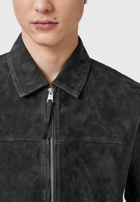 Schwarze Wildlederjacke mit Kragen und Reißverschluss vorne, mit glatter Textur und minimalen Nähten. Silberne Reißverschlussbeschläge am Verschluss.