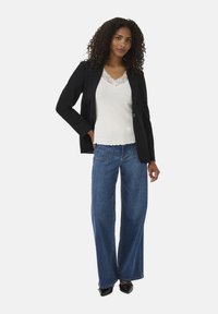 Black blazer over a white lace-trimmed top, paired with wide-leg blue jeans and black shoes. Natural, curly hair frames the face.