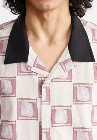 Short-sleeve shirt featuring a cream fabric with a repeating purple graphic pattern and a black collar, highlighting button closure details.