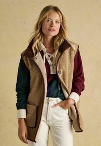 Beige Fleeceweste mit braunem Cordkragen, seitlichen Taschen und Knopfverschluss. Getragen über einem mehrfarbigen Oberteil und weißen Hosen.