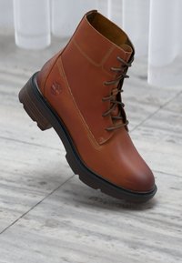 Bota de tobillo de cuero marrón con cordones, que cuenta con una suela de goma, detalles de costura y un emblema de logo en el costado. Acabado texturizado y puntera redondeada.