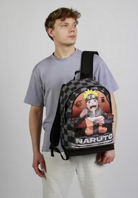 Zwarte en grijze geblokte rugzak met een kleurrafische Naruto-afbeelding aan de voorkant, voorzien van een ritsvak en verstelbare schouderbanden.