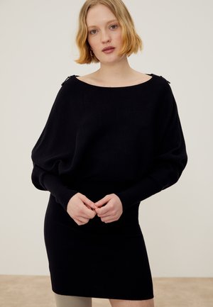 Γυναίκα με κοντά ξανθά μαλλιά που φοράει ένα μαύρο ριπ πλεκτό φόρεμα με μακριά φουσκωτά μανίκια και λεπτομέρειες φερμουάρ στους ώμους.