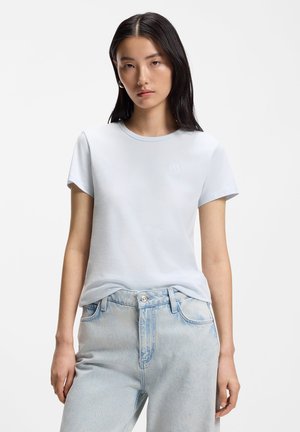 Młoda kobieta o prostych czarnych włosach ubrana w jasnoniebieski t-shirt z krótkim rękawem i jasnoniebieskie jeansy z wysokim stanem, stoi na białym tle.