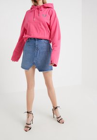 Sudadera corta rosa con bolsillo frontal y logo gris, combinada con una minifalda de mezclilla azul con un dobladillo irregular, llevada con tacones negros con tiras.