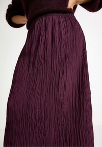 Kvinde iført en krøllet, mørkelilla maxi-nederdel med elastisk talje og matchende fluffy sweater, hænderne hvilende på hoftene.