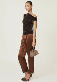 Bruine fluwelen top met één schouder en plooien, gecombineerd met glanzende bruine satijnen broeken. Accessoires bestaan uit een handtas met textuur en gouden accenten en sprankelende hakken.