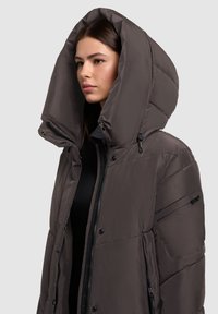 Vrouw in een donkerbruin gewatteerd jasje met een grote capuchon, staand tegen een effen lichtgrijze achtergrond, met het gezicht naar links.