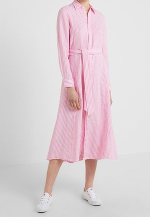 Mujer con vestido camisero rosa y blanco de rayas verticales, con cinturón y manga larga, combinado con zapatillas blancas, de pie en interiores.