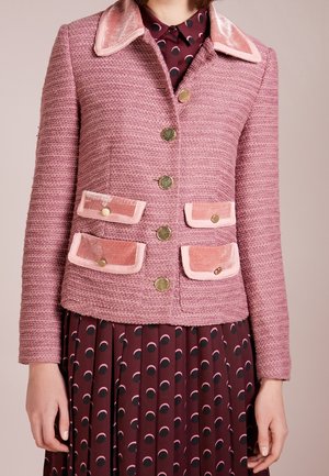 Vrouw draagt een getextureerd roze jasje met een fluwelen kraag en zakken, gouden knopen, over een bordeauxrode jurk met roze en zwarte polka dots.