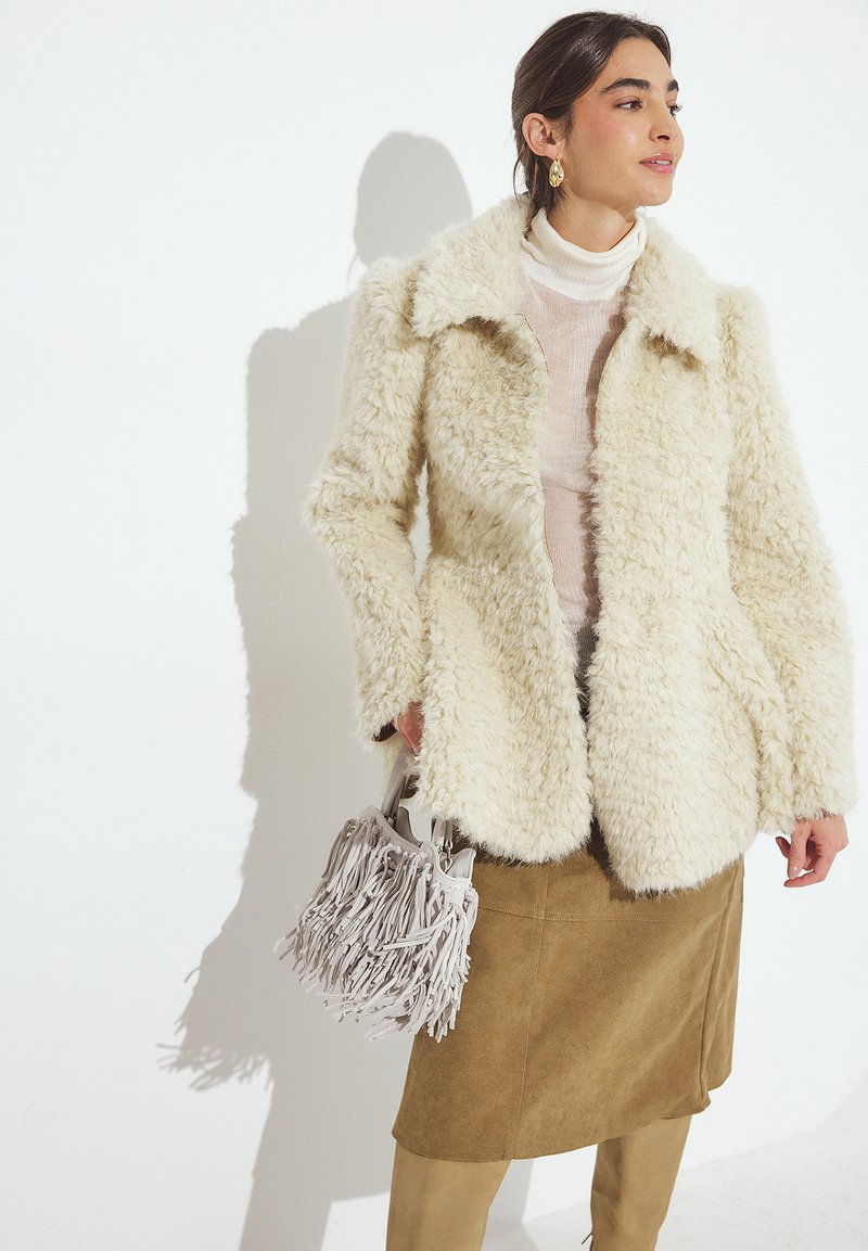 Creme farvet faux-fur jakke med krave, lagdelt over en lys turtleneck. Parret med en tan mocka nederdel og en grå frynset håndtaske.